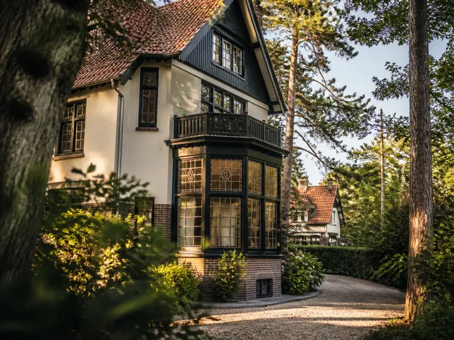 Vooroorlogse villa met houten erker en glas-in-lood, tussen hoge bomen in het late middaglicht
